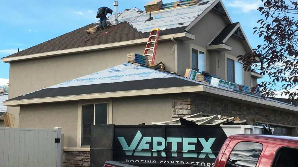 A roofer works on a house with a partially shingled roof next to a Vertex Roofing Contractors truck in West Jordan.