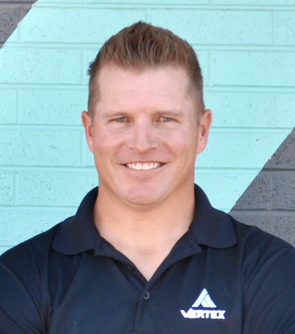 Smiling person with short hair wearing a black Vertex polo shirt stands against a geometric-patterned brick wall.