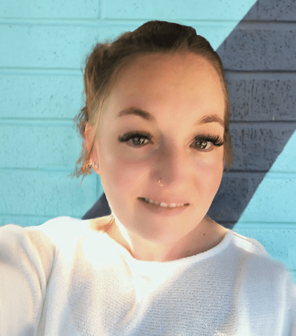 A person with tied-back hair and a nose ring smiles at the camera, standing against a brick wall painted in blue and gray diagonal sections.