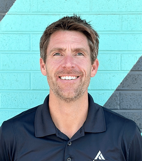 Smiling man with short hair in a black polo stands in front of a teal and black brick wall.