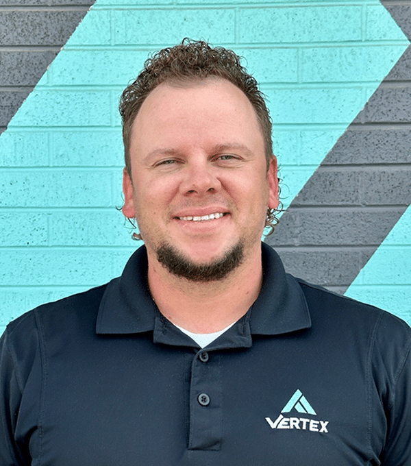 Man with short curly hair and a beard, wearing a black polo shirt with a logo, stands in front of a teal and gray brick wall.