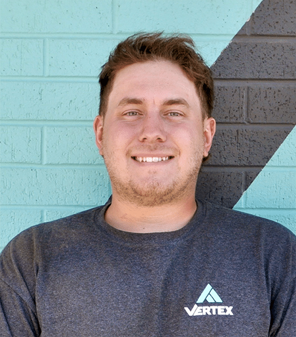 A person wearing a gray Vertex t-shirt stands in front of a geometric-patterned brick wall.