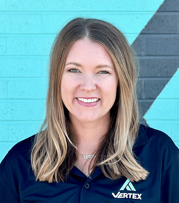 Smiling individual with long hair wearing a dark shirt with a "Vertex" logo, standing against a teal and gray brick wall.