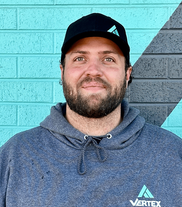 A man wearing a black cap and a gray hoodie stands against a turquoise and black brick wall.