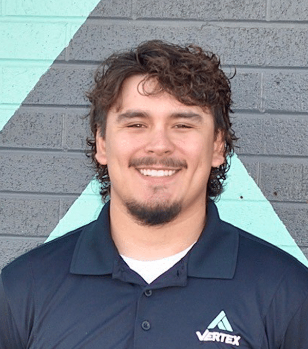 A person with a beard and curly hair smiles in front of a wall with geometric patterns. They are wearing a black polo shirt with a "VERTEX" logo.