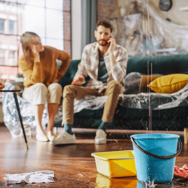 A man talks on the phone about emergency roof leak repair as water drips from the ceiling into buckets; a woman sits nearby looking concerned, hoping for a quick response in Salt Lake City.