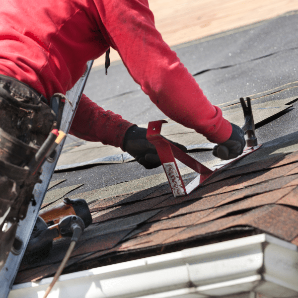 A person in a red sweatshirt installs asphalt shingles on a roof using a tool and nails, with construction equipment nearby, demonstrating the skills of a professional Salt Lake City roofing contractor.
