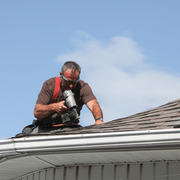A man wearing safety gear uses a nail gun to install shingles on a sloped roof under a clear sky, showcasing the expertise of a Salt Lake City roofing contractor skilled in Roof Leak Repair.