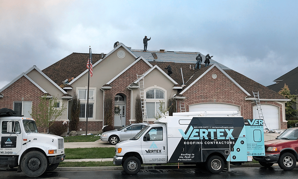 Expert roofing contractors work on a large brick house. Company vehicles, including a truck and van with "Vertex Roofing Contractors" logos, are parked in the driveway. An American flag is displayed, symbolizing commitment to quality roofing installations and repairs.