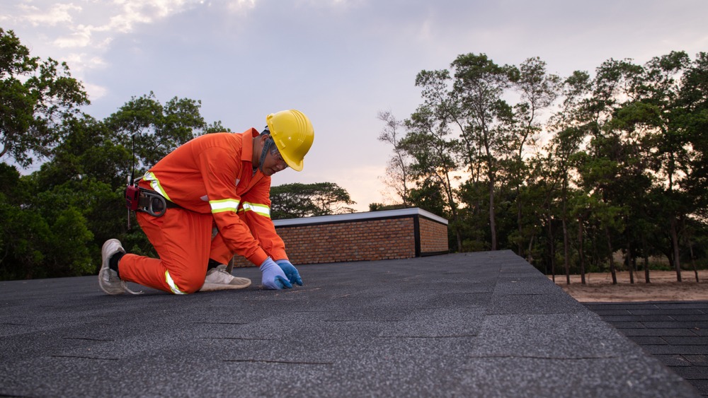 A worker in orange safety gear and a yellow hard hat kneels on a shingled roof, inspecting or repairing the surface with trees visible in the background.