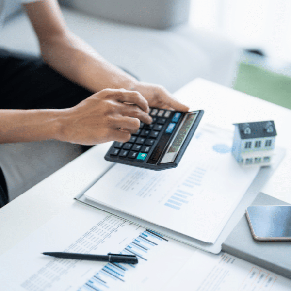 A person uses a calculator over financial documents on a table, with a small model house, a pen, and a smartphone nearby—perhaps estimating roofing costs or figuring out the cost to reroof a house.