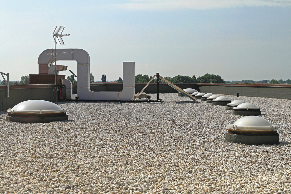 Flat gravel rooftop with several dome-shaped skylights, ventilation ducts, and an antenna—showcasing quality flat roofing installation in Utah. Trees are visible in the distance under a clear sky.