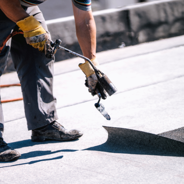 Worker in gloves uses a torch to apply and seal flat roofing materials on a flat roof surface.