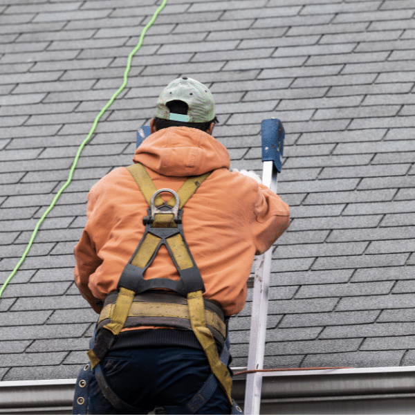 A person wearing a safety harness and orange hoodie climbs a ladder to work on a gray shingled roof, highlighting the careful process involved in roof replacement and its impact on reroofing cost.