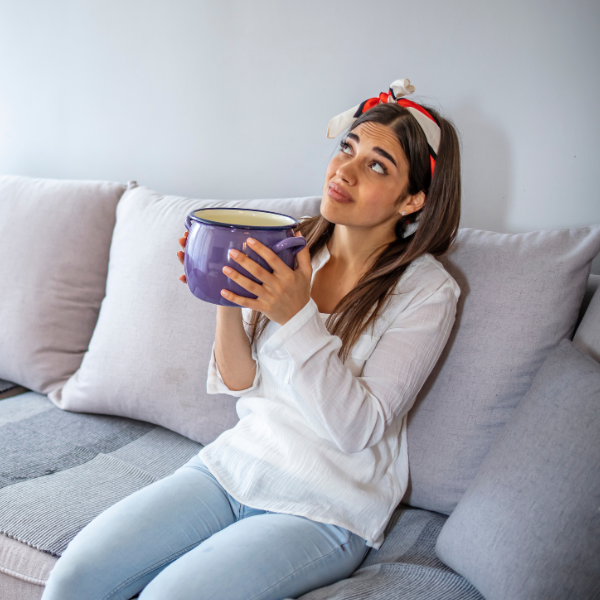 A woman sitting on a couch holds a large purple pot and looks upward with a concerned expression, perhaps worrying about reroofing costs after a leak.