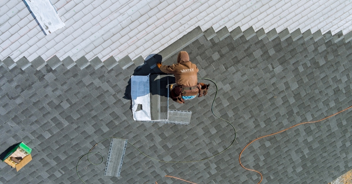 A person in a brown hoodie, likely a roofing contractor, installs or repairs shingles to address a roof leak repair on a house in Salt Lake City, with tools and materials placed nearby.