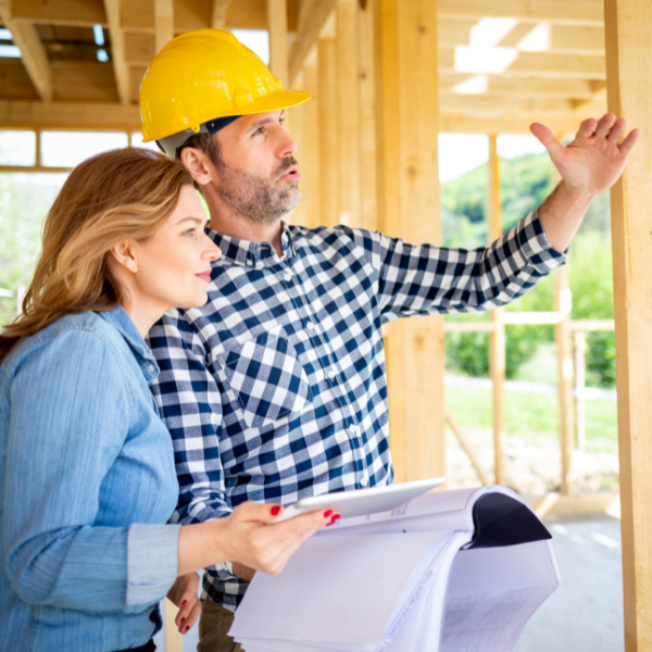 Two people at a construction site in Utah, one in a hard hat gesturing while the other holds documents and a tablet, both observing the building framework and discussing possible metal roof repair solutions.