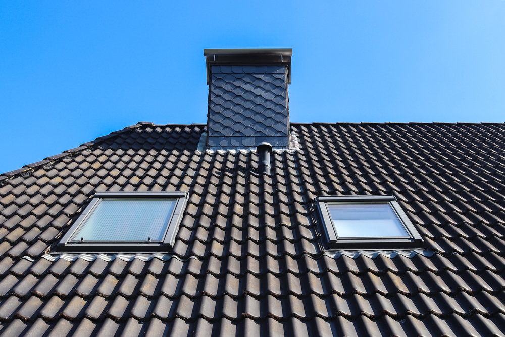 A dark tiled roof with two skylights and a shingled chimney, illustrating a professional metal roof installation against a clear blue sky.